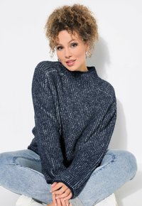 Marineblauer, gerippter Strickpullover mit hohem Kragen und breiten Bündchen, kombiniert mit hellblauen Jeans. Weiche Textur, lockere Passform und leichter Glanz.