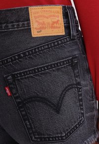 Pantalones vaqueros negros de mezclilla con un dobladillo deshilachado, con un parche de cuero color canela con el logo y una etiqueta roja en el bolsillo trasero. Detalles de textura desgastada visibles.