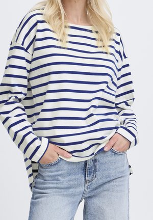 En blond person iført en hvid og marineblå vandret striped langærmet skjorte og lyseblå vaskede jeans med hænderne i lommerne.