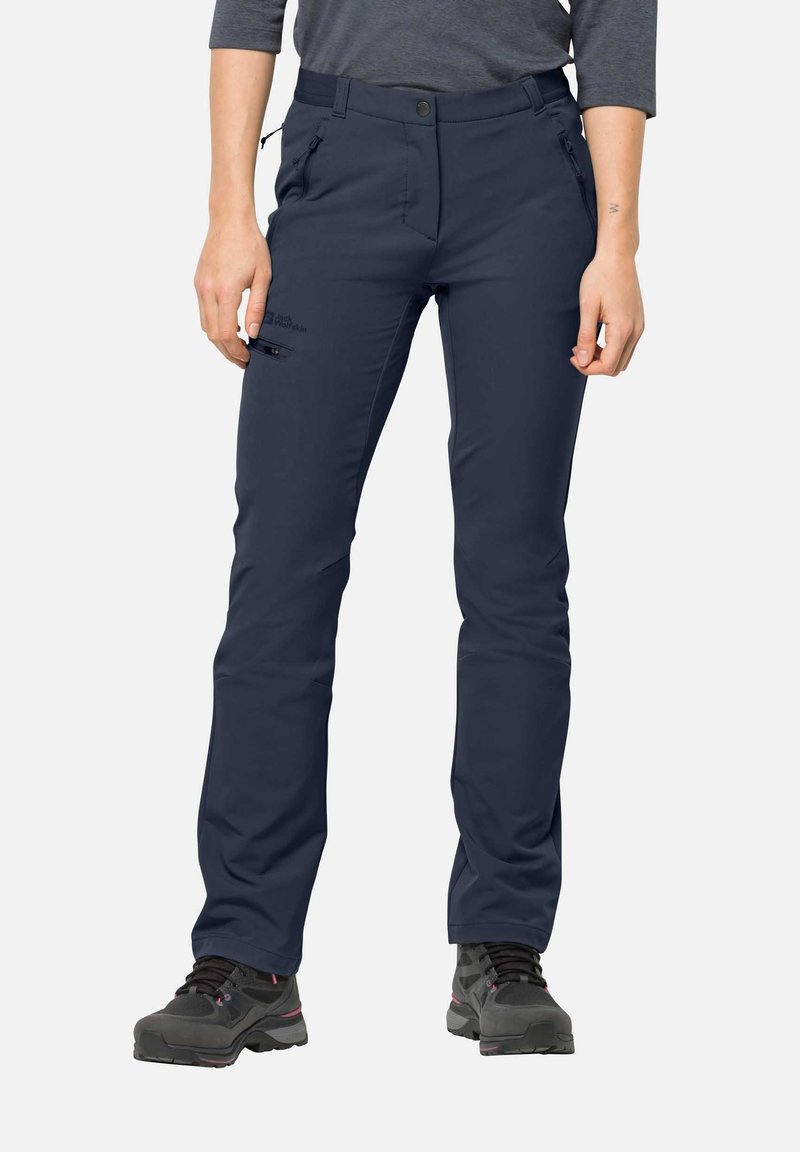 Personne portant un pantalon d'extérieur bleu marine avec des poches zippées et des chaussures de randonnée foncées, debout devant un fond uni.