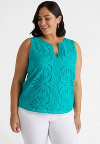 Top senza maniche di colore teal con intricate aperture decorative, scollatura a V e vestibilità ampia. Abbinato a pantaloni bianchi per un contrasto.