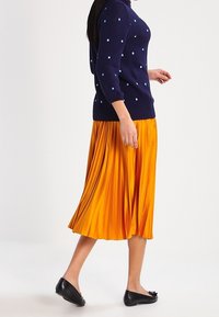 Marinblå tröja med ljusblåa prickar, ihopparad med en plisserad, knälång, klar gul kjol och svarta lägenheter.