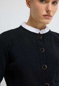 Musta vaate, jossa on teksturoidut kankaat, korkea röyhelötty valkoinen kaulus ja kultasävytteiset nappikoristeet edessä.