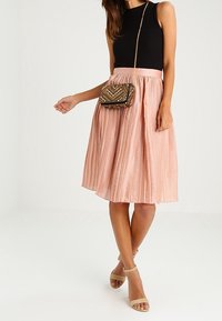 Donna che indossa un top senza maniche nero, una gonna midi plissettata rosa, sandali beige con cinturino alla caviglia e una piccola borsa a tracolla con motivo.