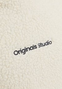 Tessuto in pile di colore crema con una superficie testurizzata e soffice. Presenta il testo ricamato in nero "Originals Studio." Nessun'altra decorazione.