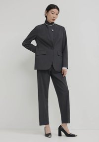 Completo grigio su misura con blazer a un bottone e pantaloni abbinati. Scarpe con tacco alto nere e gioielli in argento completano il look.