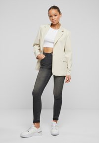 Blazer beige con rever a punta, jeans skinny neri a vita alta e sneaker bianche. Il blazer ha tasche frontali e una vestibilità morbida.