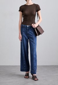 Brauner strukturierter Oberteil, weit geschnittene blaue Jeans mit Seitennähten und braunen Sandalen mit Absatz. Braune Umhängetasche quer über den Körper getragen.