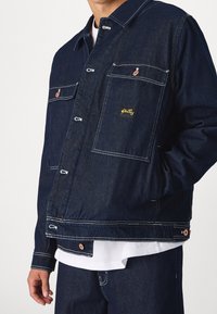 Tmavě modrá džínová bunda s dvěma náprsními kapsami a jednou boční kapsou. Má bílé švy a malou žlutou značku na hrudi.