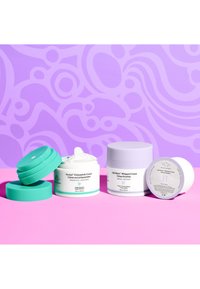 Four skincare cream containers on a pink surface. Includes a green-lidded jar, a light purple jar, and two white jars with labels.