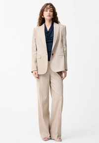 Femme debout portant un costume beige à fines rayures avec un pantalon à jambes larges et un blazer par-dessus un top drapé bleu marine et des talons ouverts.