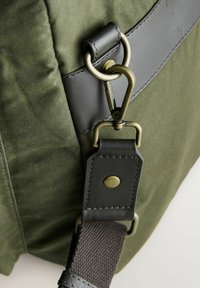 Olajzöld textil táska fekete bőrszárnyakkal, bronz fém kapoccsal, texturált pánttal és gombos bőrfelülettel.