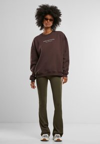 Hnědá oversized mikina s grafickým textem, v kombinaci se zelenými žebrovanými rozšířenými kalhotami. Modelka má na sobě světlé tenisky a sluneční brýle.