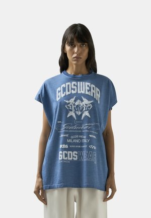 Donna che indossa una maglia senza maniche oversize blu GCDSWEAR con testo grafico bianco e simboli, in piedi davanti a uno sfondo chiaro e uniforme.