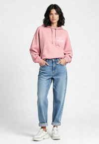 Felpa rosa con stampa "Hôtel de L'Amour", vestibilità oversize, tasca frontale. Abbinata a jeans blu rilassati e sneaker bianche.