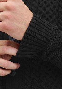 Schwarzer Strickcardigan mit gerippten Bündchen und strukturierten Mustern. Verfügt über Knöpfe an der Vorderseite und zeigt eine Nahaufnahme einer Hand, die das Bündchen anpasst.
