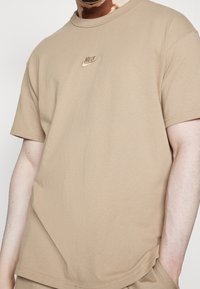 Beige bavlněné tričko s krátkými rukávy, kulatým výstřihem a malým vyšívaným logem Nike na hrudi. Hladká textura.