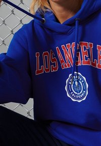 Modrá mikina s kapucí s nápisem "LOS ANGELES" červenými písmeny, s kruhovým bílým logem na hrudi. Jsou viditelné šňůrky a textura vypadá měkce.