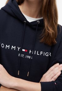 Personne portant un sweat à capuche Tommy Hilfiger bleu marine avec texte blanc et petit logo rouge, blanc et bleu sur la poitrine et la manche.