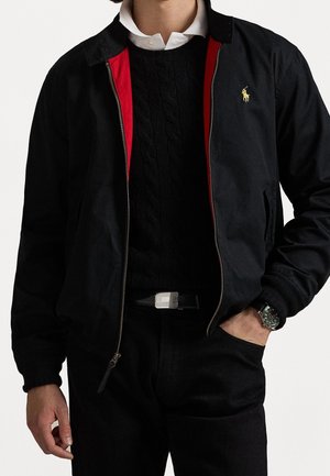 Mann trägt schwarze Reißverschlussjacke mit roter Innenfütterung über einem schwarzen Kabelstrickpullover und weißem Hemd mit Kragen, schwarze Hose, Gürtel und Armbanduhr.