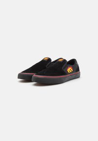 Melni slip-on apavi no zamšas, ar sarkanu akcentu joslu un izšūtu logotipu ar liesmu detaļām sānos.