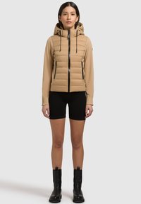 Beige puffjakke med en tætsiddende design, hætte og sort lynlås. Har ribbede manchetter og sidelommer. Båret med sorte shorts og ankelstøvler.