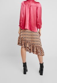 Blusa rosa lucida con maniche lunghe, abbinata a una gonna asimmetrica con volant a fantasia nei colori arancione, nero e bianco. Stivali neri alla caviglia.