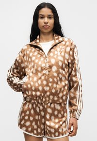 Femme portant une veste zippée marron et blanche à pois et un short assorti avec des bandes blanches sur les manches et les côtés.