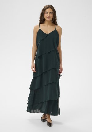 Kobieta o długich kręconych włosach, ubrana w ciemnozieloną suknię maxi z falbanami i czarne buty w szpic, stojąca na tle jednolitego tła.