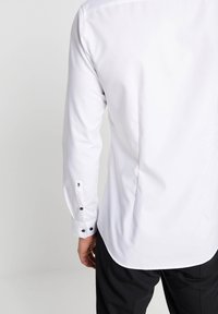 Achteraanzicht van een persoon die een nauwsluitend wit shirt met lange mouwen en afgeknopte manchetten en een zwarte broek draagt, tegen een effen achtergrond.