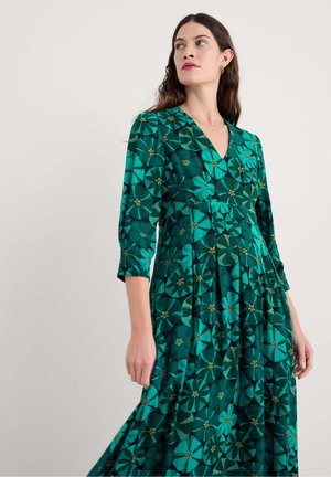 Vrouw in een groene bloemen V-halsjurk met driekwartmouwen, die naar de zijkant kijkt tegen een effen lichte achtergrond.