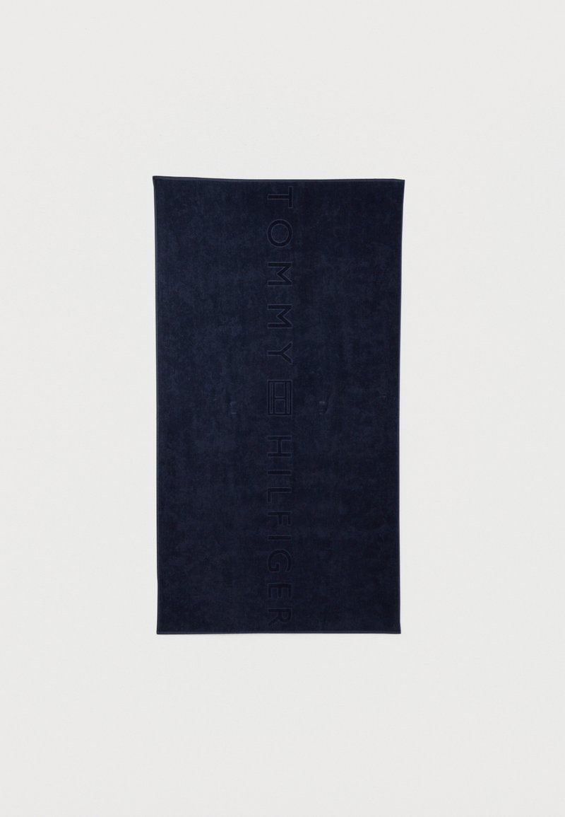 Serviette bleu marine foncé en matériau doux, avec le logo "TOMMY HILFIGER" en relief sur un côté. Forme rectangulaire, surface texturée.