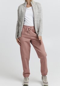 Giacca a zip in maglia grigia chiara, t-shirt bianca e pantaloni sportivi rosa pallido con vita e polsini elasticizzati, abbinati a sneakers grigie.