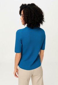 Blå ribbad stickad topp med korta ärmar, med rund halsringning och en något böjd fåll. Bärs med beiga byxor.