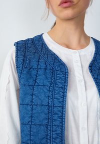 Blå vadderad väst med en strukturerad yta, med en rundad nacke och en knäppt vit skjorta under för lagerpåklädning.