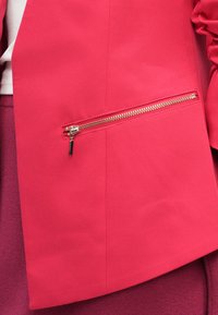 Blazer rosa acceso con una tasca a zip visibile con dettagli metallici. Texture liscia e design su misura, parte di un outfit a strati.