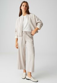 Femme portant une veste bomber beige, une chemise texturée blanche, un pantalon beige à jambes larges, et des mocassins blancs, debout devant un fond neutre.