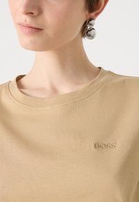 Beige katoenen t-shirt met een ronde halslijn en subtiele borduursels van het woord "BOSS." Zilveren ronde oorbellen complementeren het shirt.