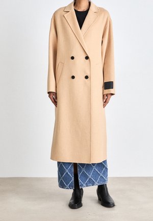 Person trägt einen beigen doppelreihigen langen Mantel, blauen gemusterten Jeansrock, schwarzes Top und schwarze Knöchelstiefel, steht auf einem grauen Boden.