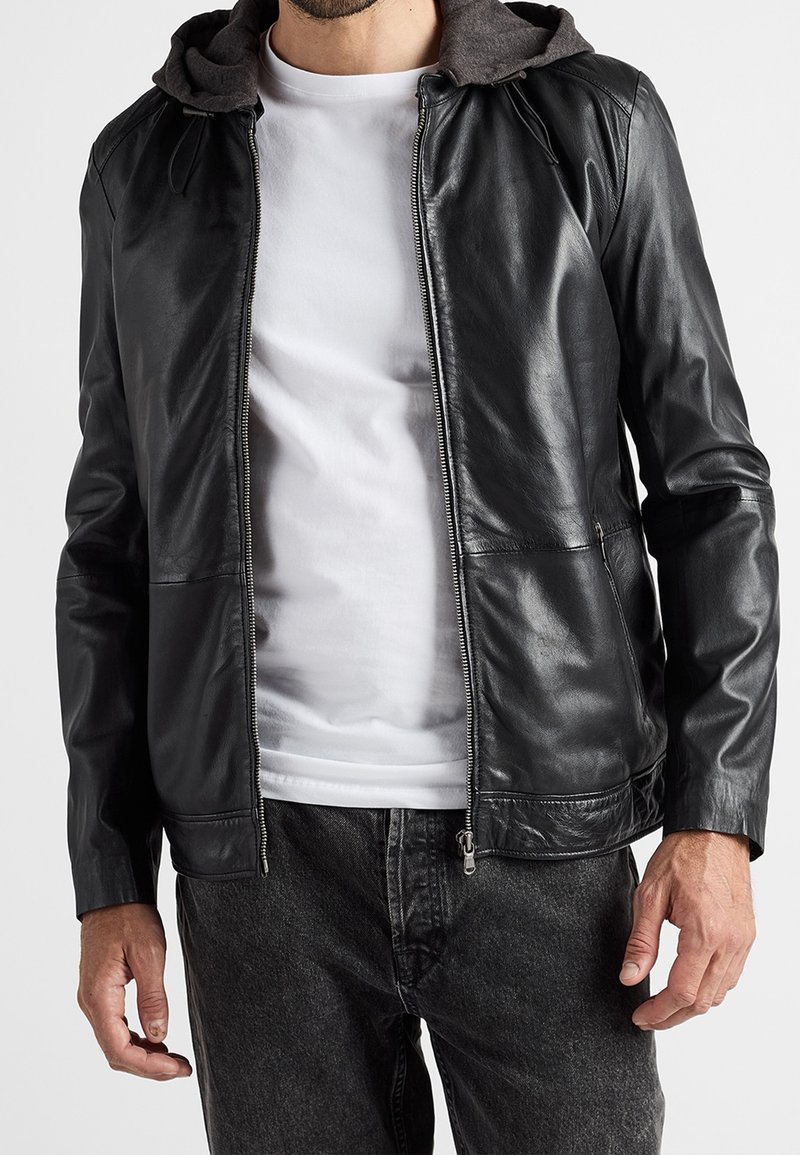 Černá kožená bunda se šedou kapucí, předním zipem a dvěma bočními kapsami. Nositelná přes bílé tričko a doplněná tmavými džínami.