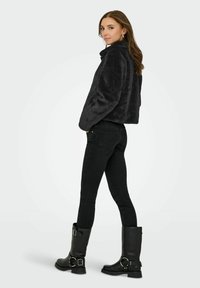 Femme portant une veste polaire noire, un jean skinny noir et des bottes noires à boucle, debout les mains dans les poches, regardant par-dessus son épaule.
