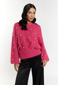 Maglione rosa fluffy con una vestibilità morbida, orlo a coste e numerose perle decorative distribuite uniformemente su tutta la stoffa. Abbinato a pantaloni neri.
