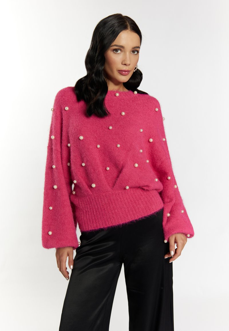 Maglione rosa fluffy con una vestibilità morbida, orlo a coste e numerose perle decorative distribuite uniformemente su tutta la stoffa. Abbinato a pantaloni neri.