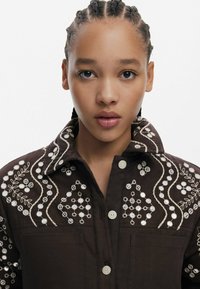 Brązowa haftowana kurtka z białymi koralikami i skomplikowanymi wzorami kwiatowymi, z usztywnionym kołnierzem i zapięciem na guziki z przodu.