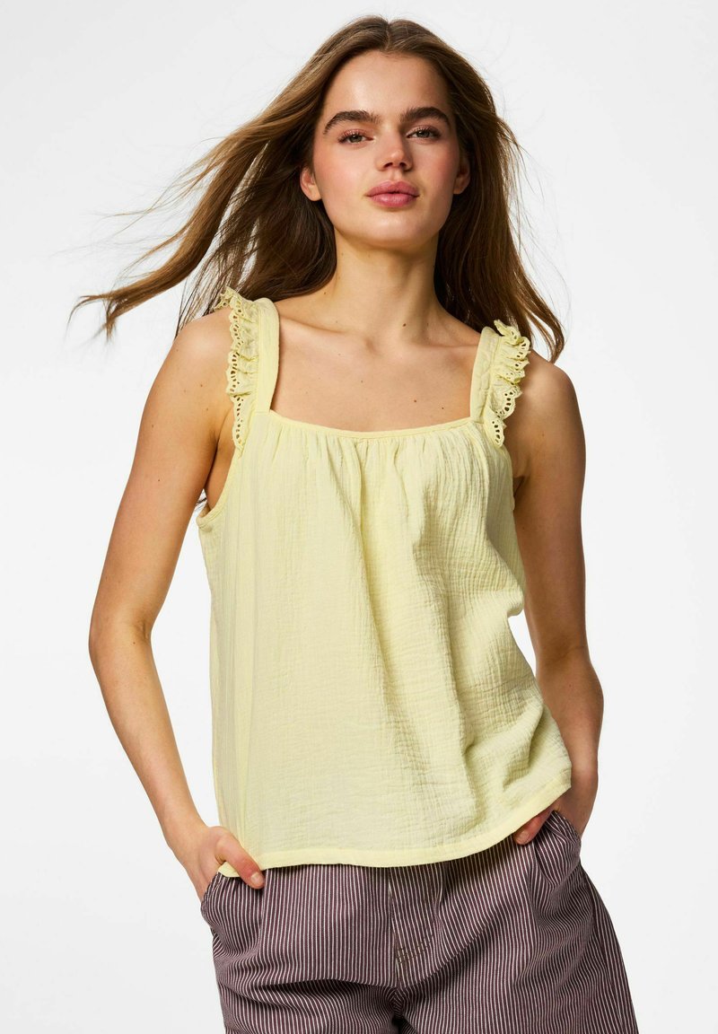 Jeune femme aux longs cheveux portant un haut sans manches jaune clair avec des bretelles volantées et un pantalon rayé, mains dans les poches, fond blanc.