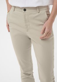 Pantalons beiges en tissu lisse, avec une coupe slim, fermeture à bouton et poches latérales. Détails de couture subtils visibles.