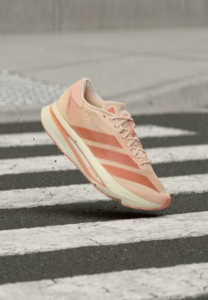 Zapatilla de correr Adidas en tonos rosa claro y coral en el aire sobre un paso de peatones blanco y negro con pequeños escombros volando debajo.