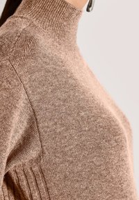 Nahaufnahme einer Person, die einen hellbraunen, gestrickten Rollkragenpullover mit geripptem Kragen und seitlichen Rippendetails trägt.