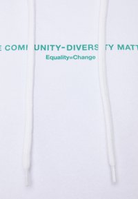 Felpa bianca con una texture morbida. Presenta un testo stampato in verde: "LA COMUNITÀ - LA DIVERSITÀ È IMPORTANTE Uguaglianza = Cambiamento." Inclusi i lacci.