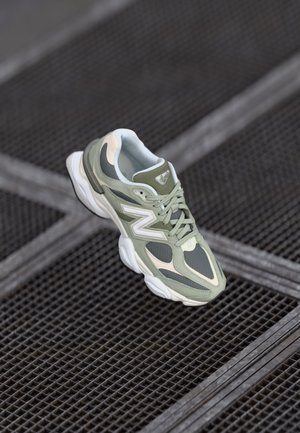 Gray and green New Balance sneaker with white sole resting on black metal grates arranged in a grid pattern.